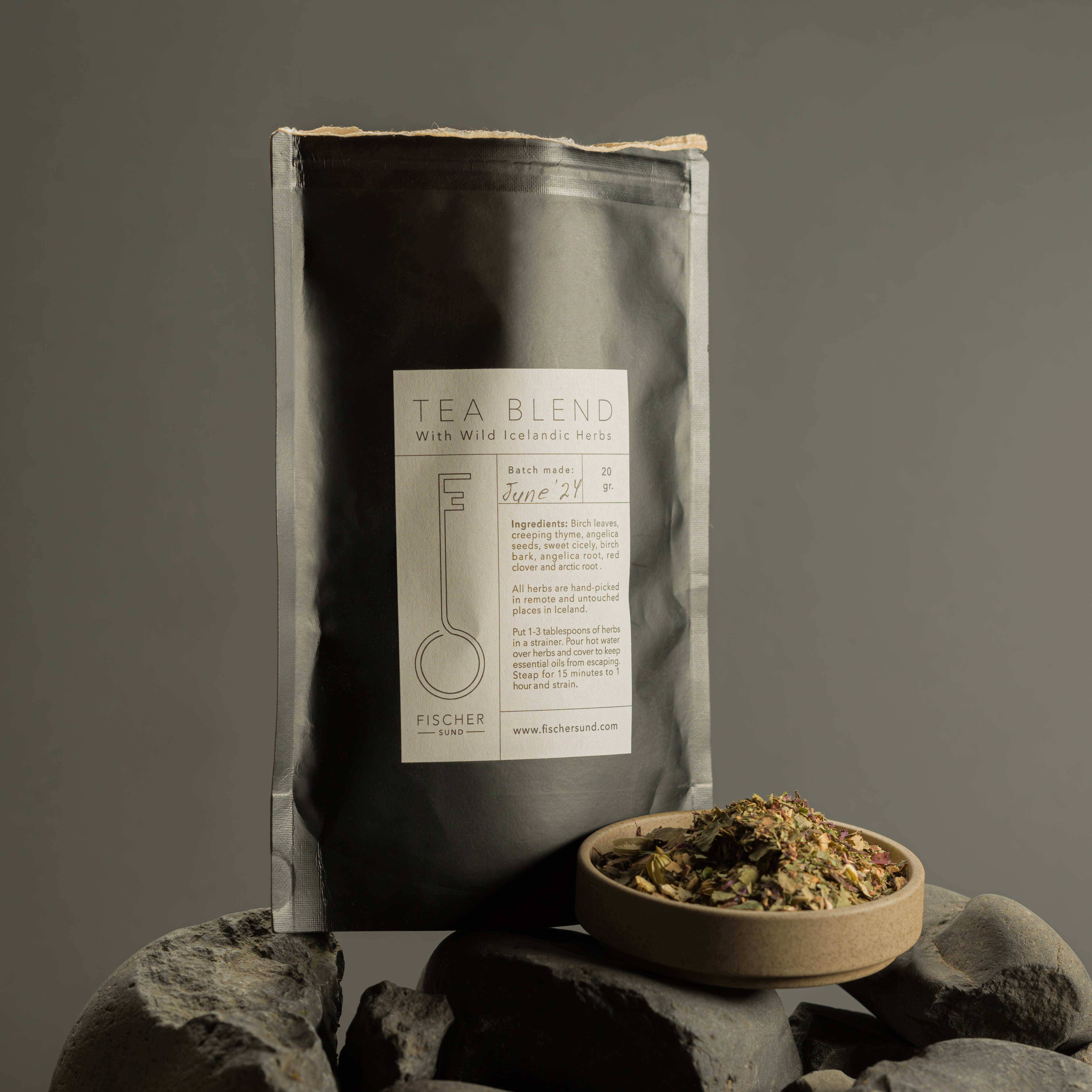 A Fischersund black tea bag labeled 'Tea Blend' with a graphic of a teapot, resting on a rocky surface with a small dried herb mix beside it.